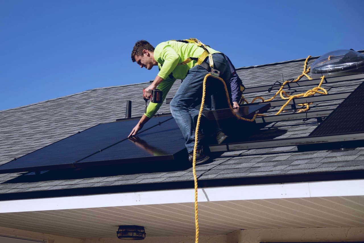 Solar installer in a safety harness fitting panels on a shingle roof